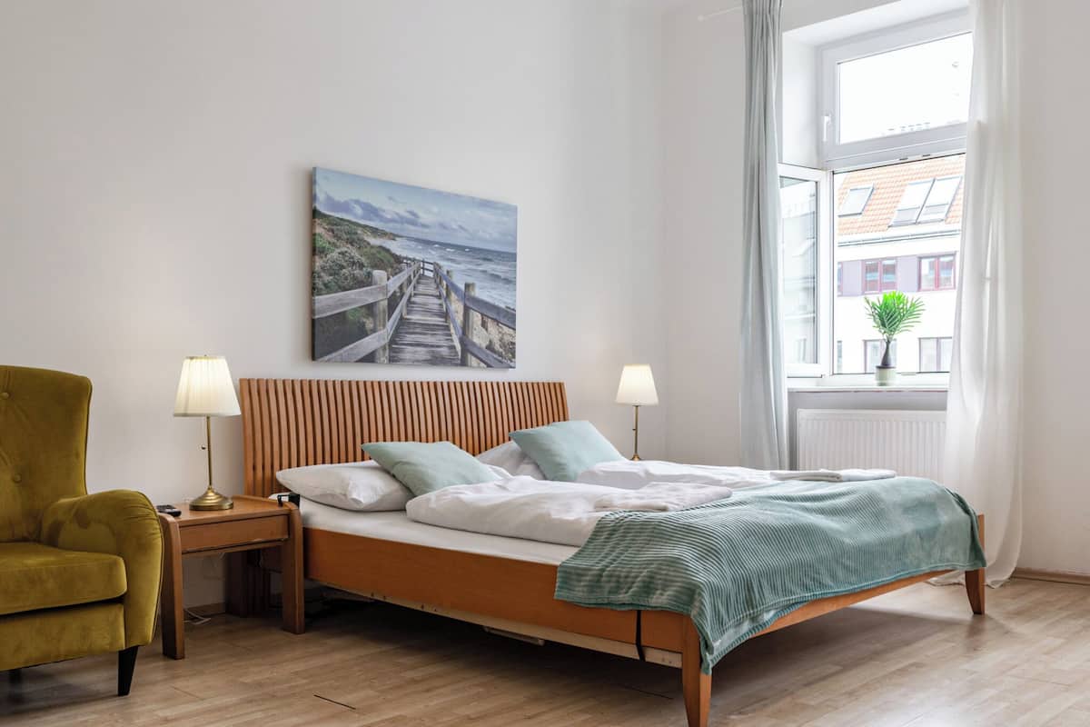 Een slaapkamer met een houten bed, twee bedlampen, een groene plaid, een schilderij van een kustlandschap, een raam met gordijnen en een kleine plant op de vensterbank.