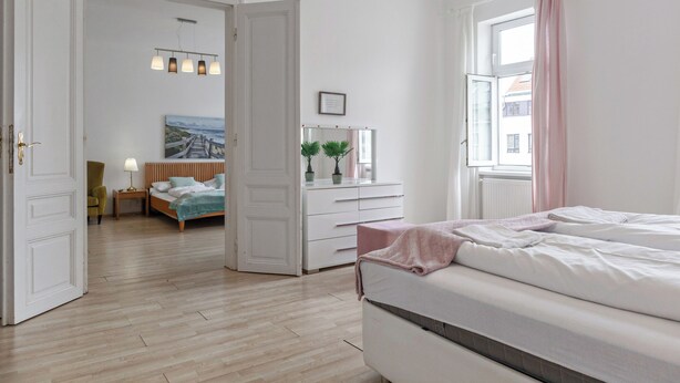 A hotel room with a wooden floor, a large bed, a bedside table, a dresser, a window with curtains, and a hallway leading to another room. - Elegant Room Shared Apt Heart of Vienna (Vienna)