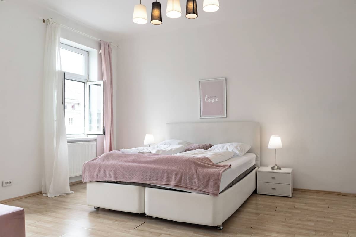 Een slaapkamer met een bed, nachtkastjes, een raam met gordijnen, een plafondlamp en een lijst met een afbeelding aan de muur.