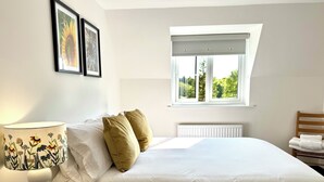 Quarto com cama, mesa de cabeceira com luminária, janela com vista para árvores e cadeira com toalhas.