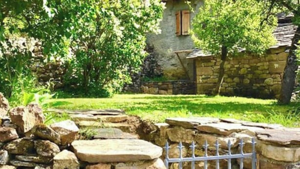 Una cabaña de piedra con un muro de piedra y una reja metálica, rodeada de abundante vegetación.