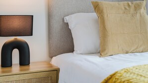 Cama bem arrumada com roupa de cama branca e travesseiro bege, luminária de cabeceira e mesa de cabeceira de madeira com gaveta trançada.