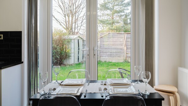 Mesa de jantar posta para dois com vista para um jardim através de uma porta de correr de vidro.