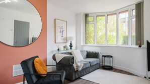 Sala de estar com sofá cinza, uma cadeira preta, um espelho redondo e uma janela com cortinas.