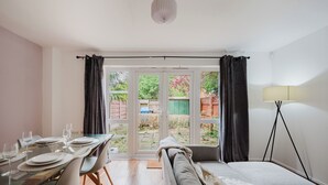 Una habitación con una mesa de comedor lista para cuatro personas, un sofá, una lámpara y vistas a un jardín a través de una puerta de cristal.