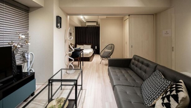 A modern living room with a sofa, a glass coffee table, and a TV on a stand. There is a chair and a bed in the background. - Mondomio Sapporo 207 (Sapporo)