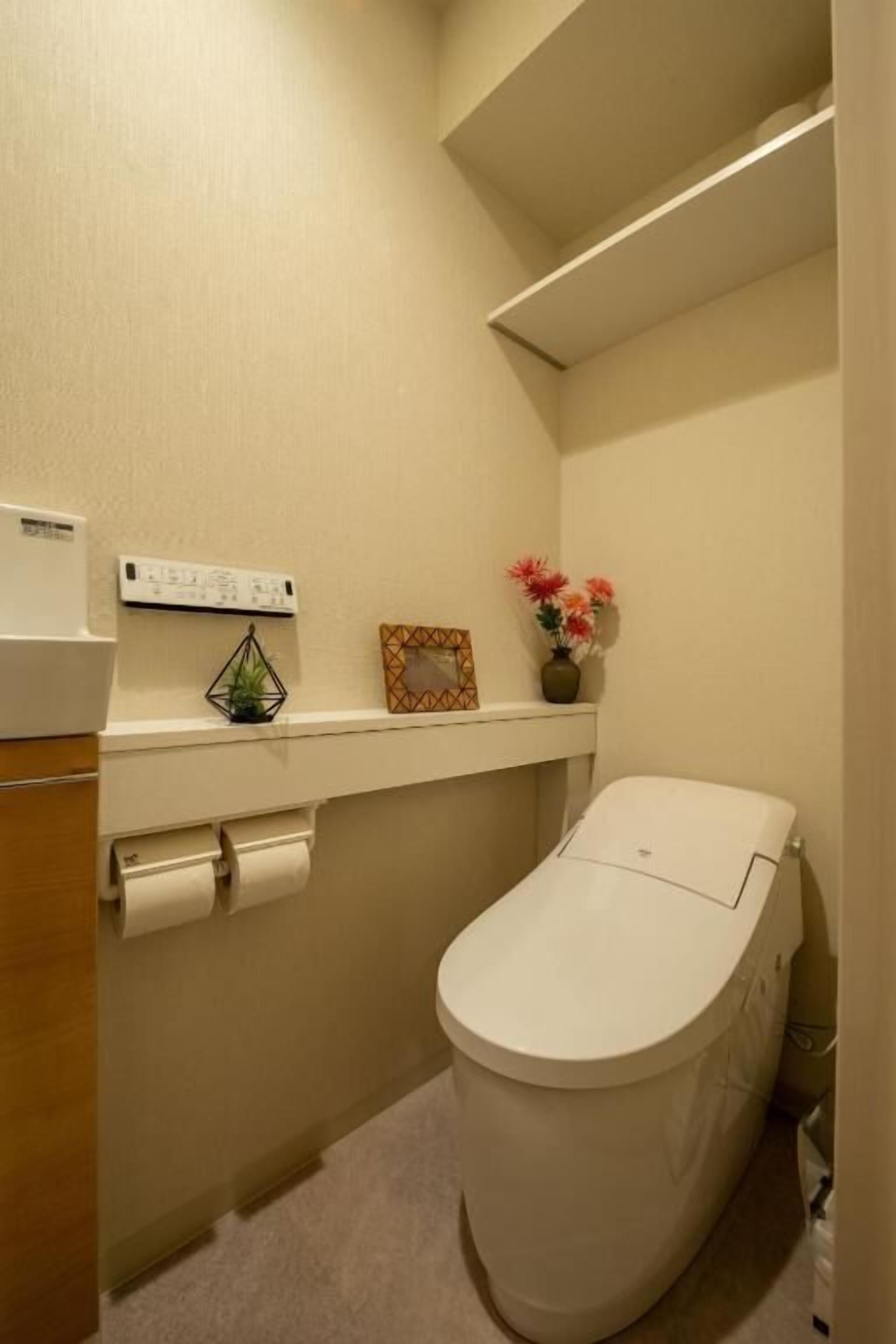 Une petite salle de bain comprenant des toilettes, une étagère avec une petite plante et un cadre photo, ainsi qu’un porte-rouleau de papier toilette.