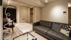 A modern hotel room with a bed, a grey sofa, and a glass coffee table. There is a wardrobe with a mirror and a coat rack. - Mondomio Sapporo 207 (Sapporo)