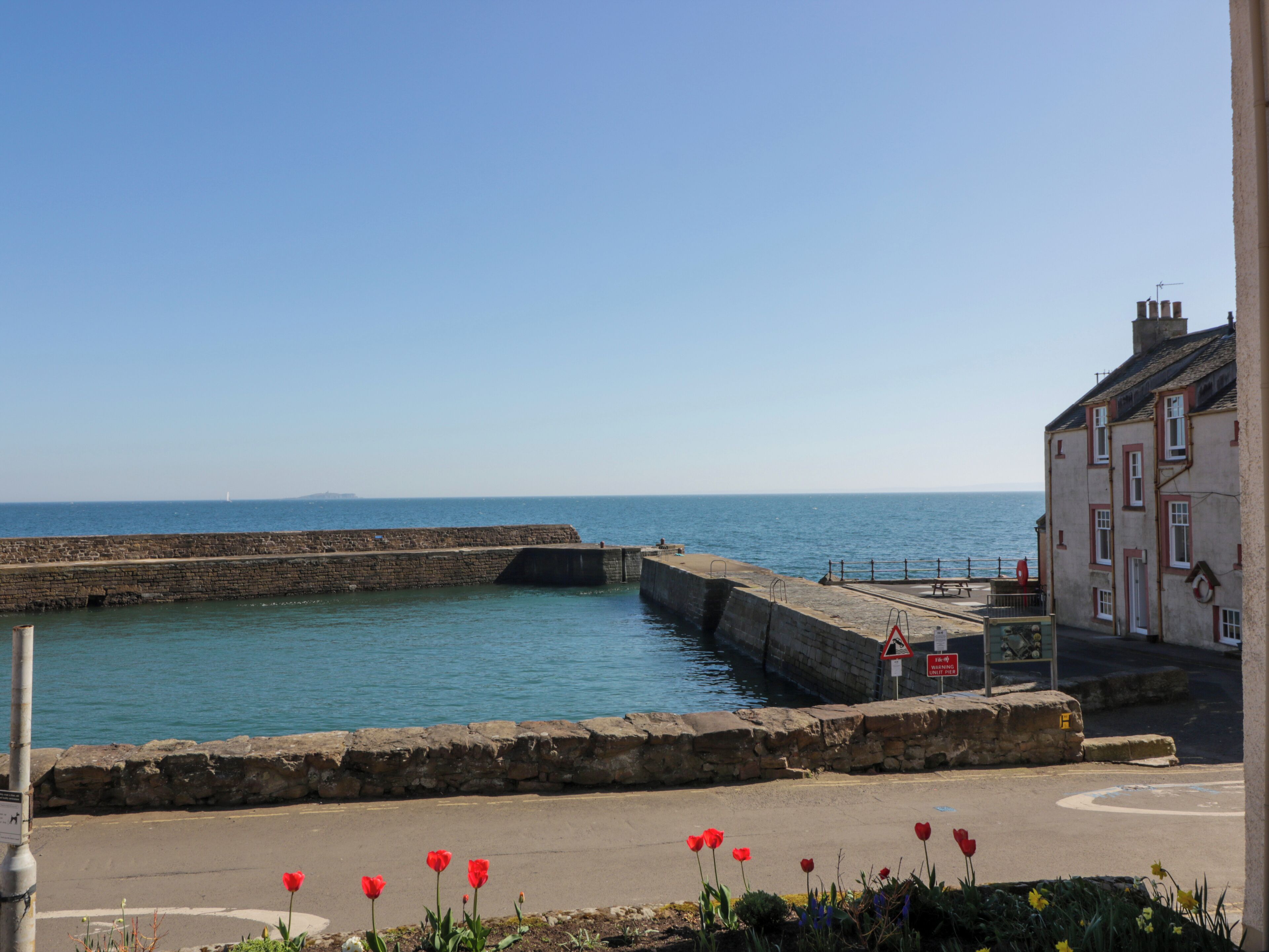 A coastal view with a stone quay, a clear sky, and a building with a red door.