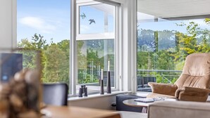 A modern living room with a large window, a comfortable sofa, a small table, and a balcony with a view of greenery.