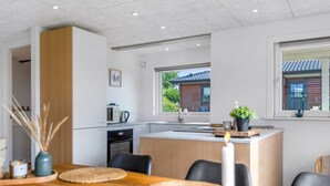 A modern kitchen with a central island, a dining area with a wooden table and chairs, and a view of a traditional house through the window.
