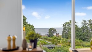 A view from a room with a large window overlooking a body of water, a garden, and a small building.
