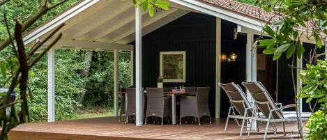 A covered outdoor seating area with a table and chairs, surrounded by greenery.