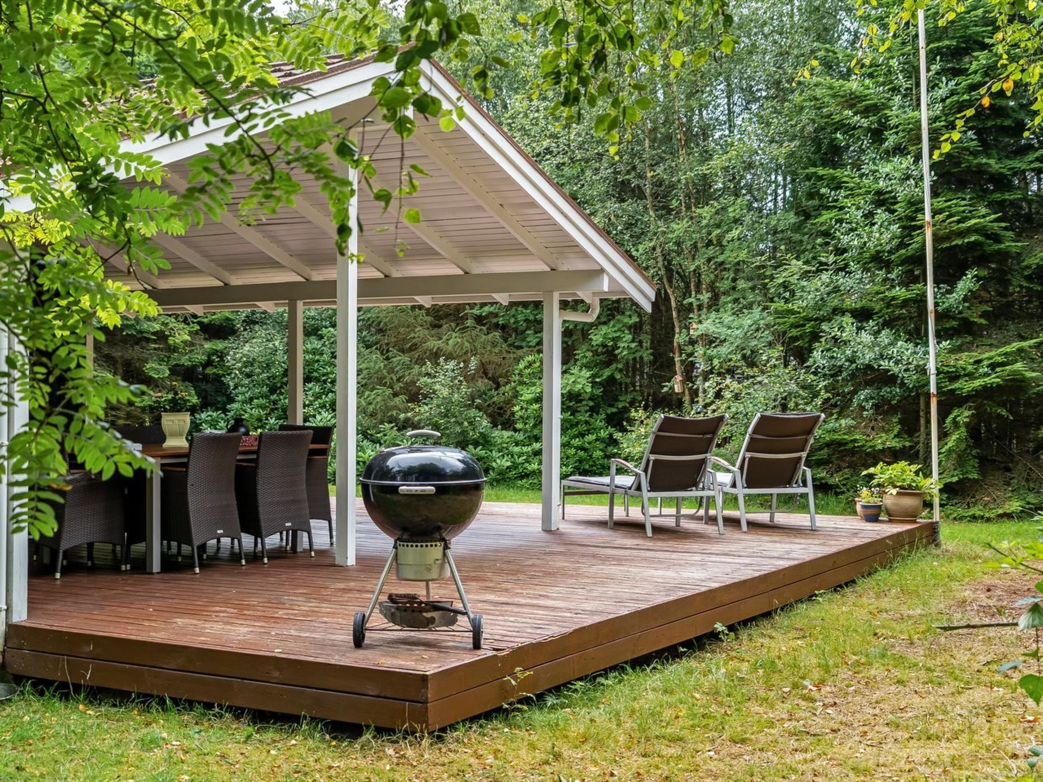 Un patio cubierto con una parrilla, mobiliario exterior y abundante vegetación.