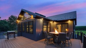 Una terraza moderna al aire libre con un hogar para fuego y una zona de estar, rodeada por una cubierta de madera y vistas al campo.