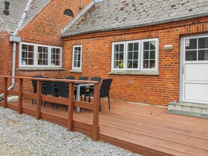 Un bâtiment en briques avec une terrasse en bois, une table entourée de chaises et une porte blanche.