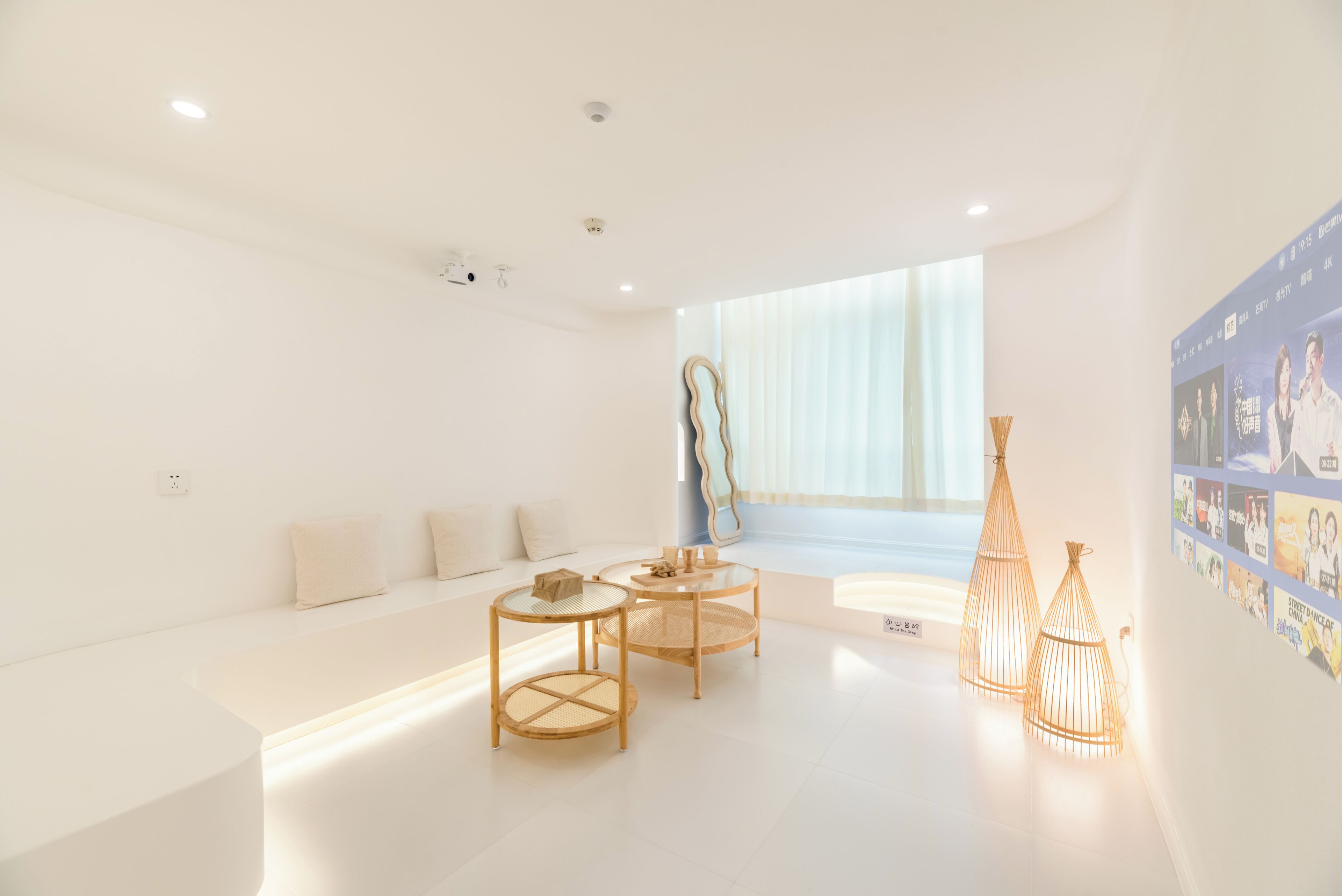 A modern, minimalist room with a white sofa, two round tables with woven tops, and a large window with sheer curtains.