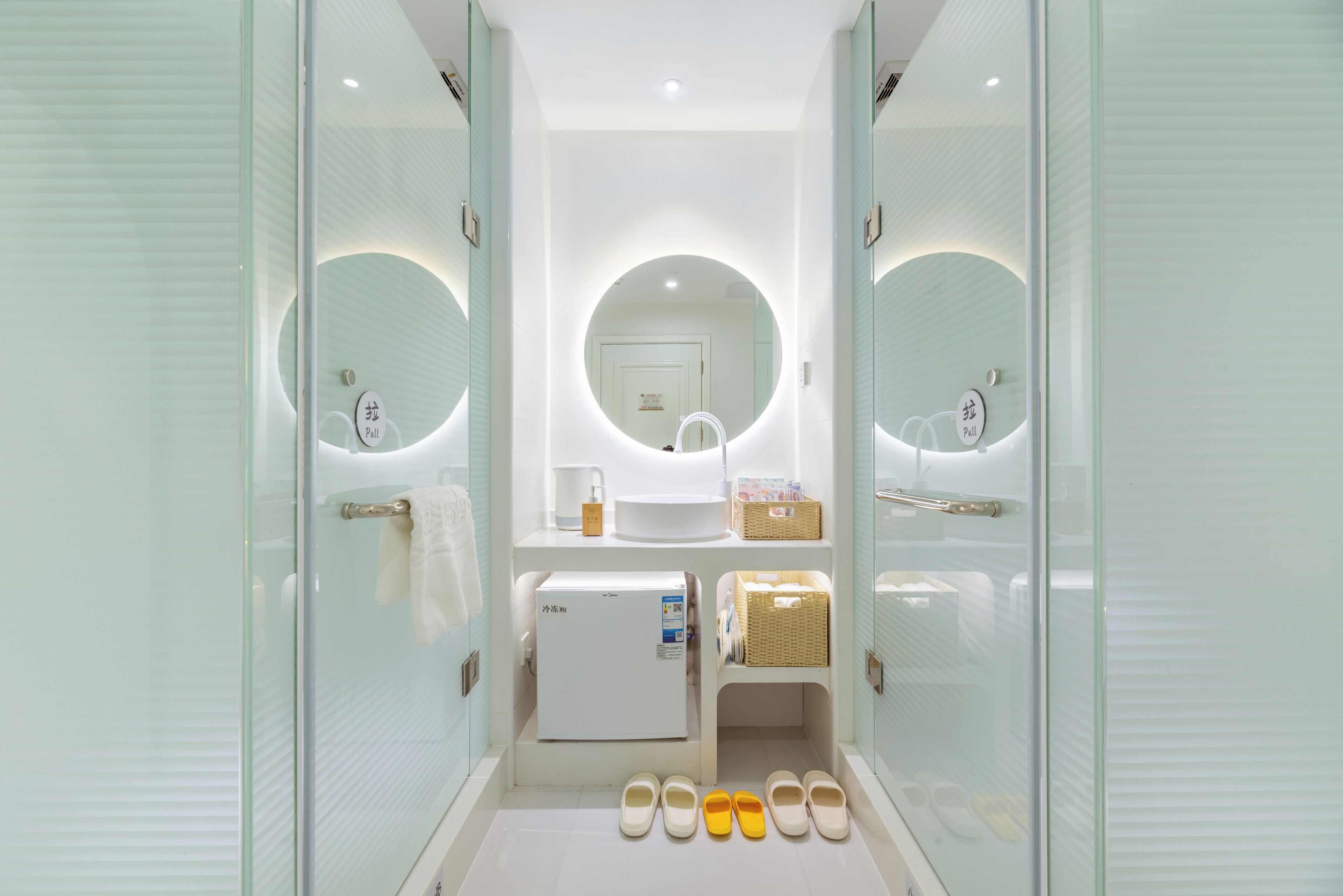 A modern bathroom with a glass partition, a round mirror, a small refrigerator, and a shelf with toiletries.