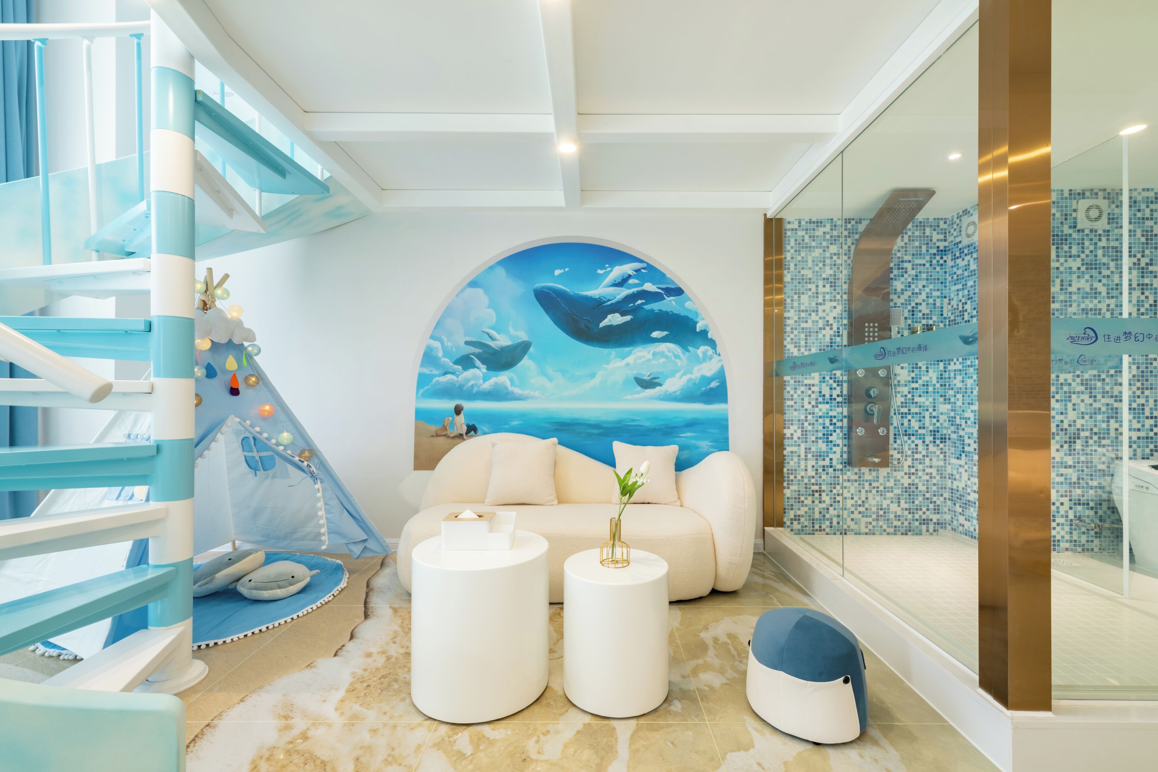 Une chambre d’hôtel moderne aux couleurs bleu et blanc, avec une fenêtre ronde ornée d’une fresque représentant une baleine, un canapé, deux tables rondes et une douche en verre.