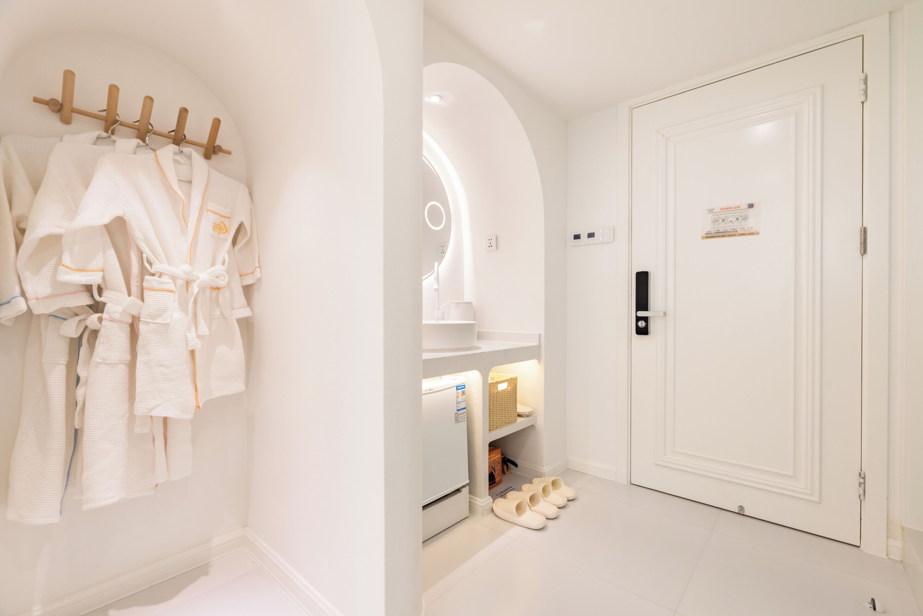 A hotel room with a white robe rack, a refrigerator, slippers, and a door with a sign.