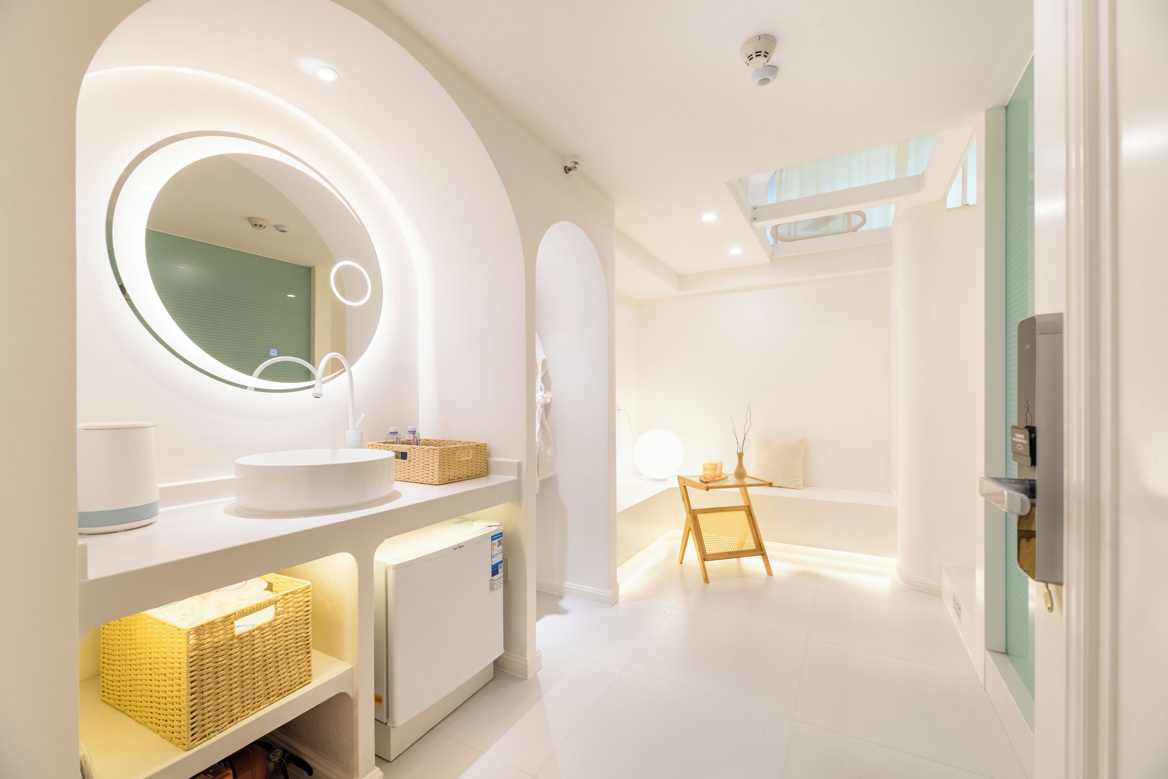 A modern bathroom with a round mirror, white sink, and a small refrigerator.