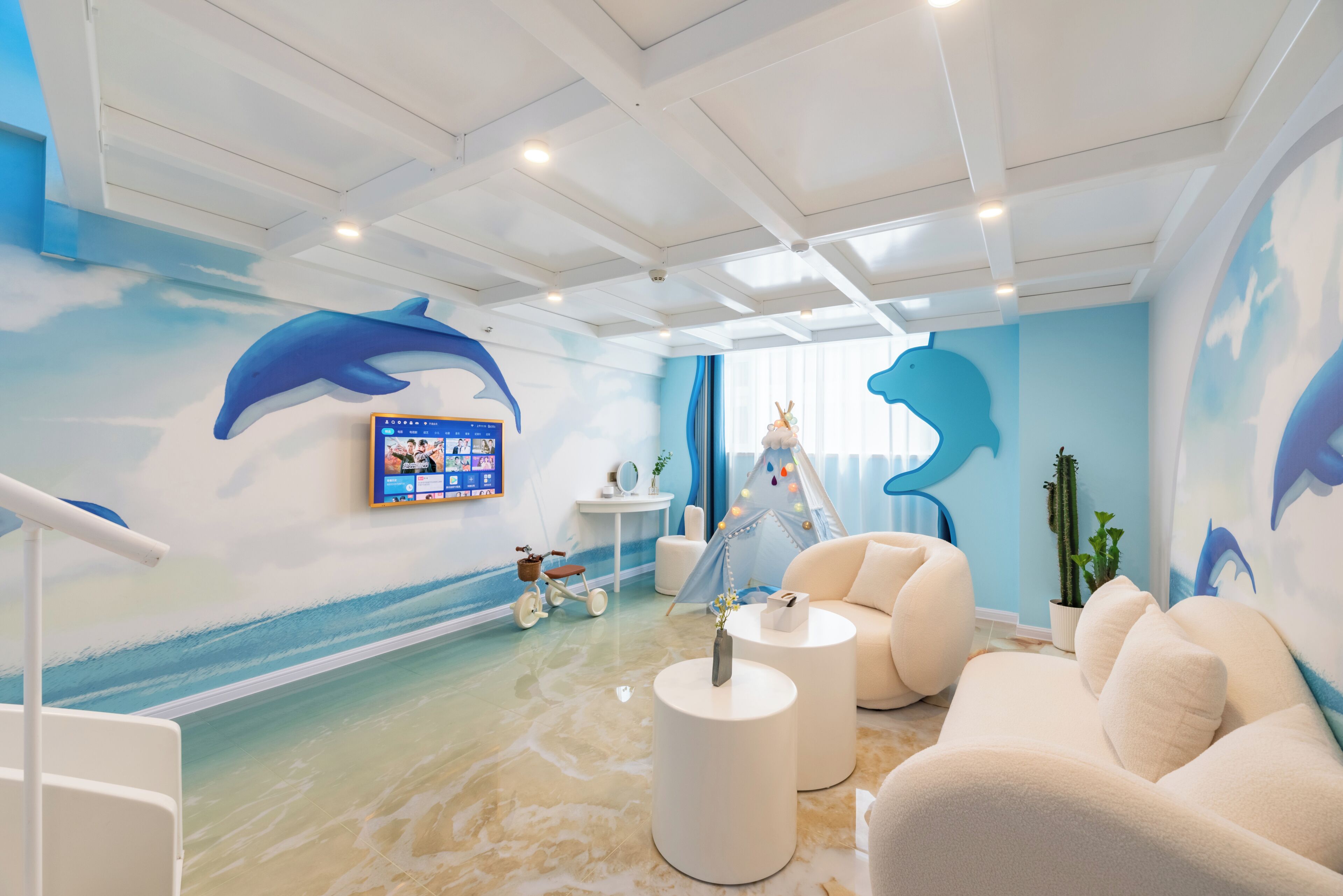 Una habitación con temática playera, que presenta un mural grande de delfines, una tienda de campaña y un televisor en la pared.