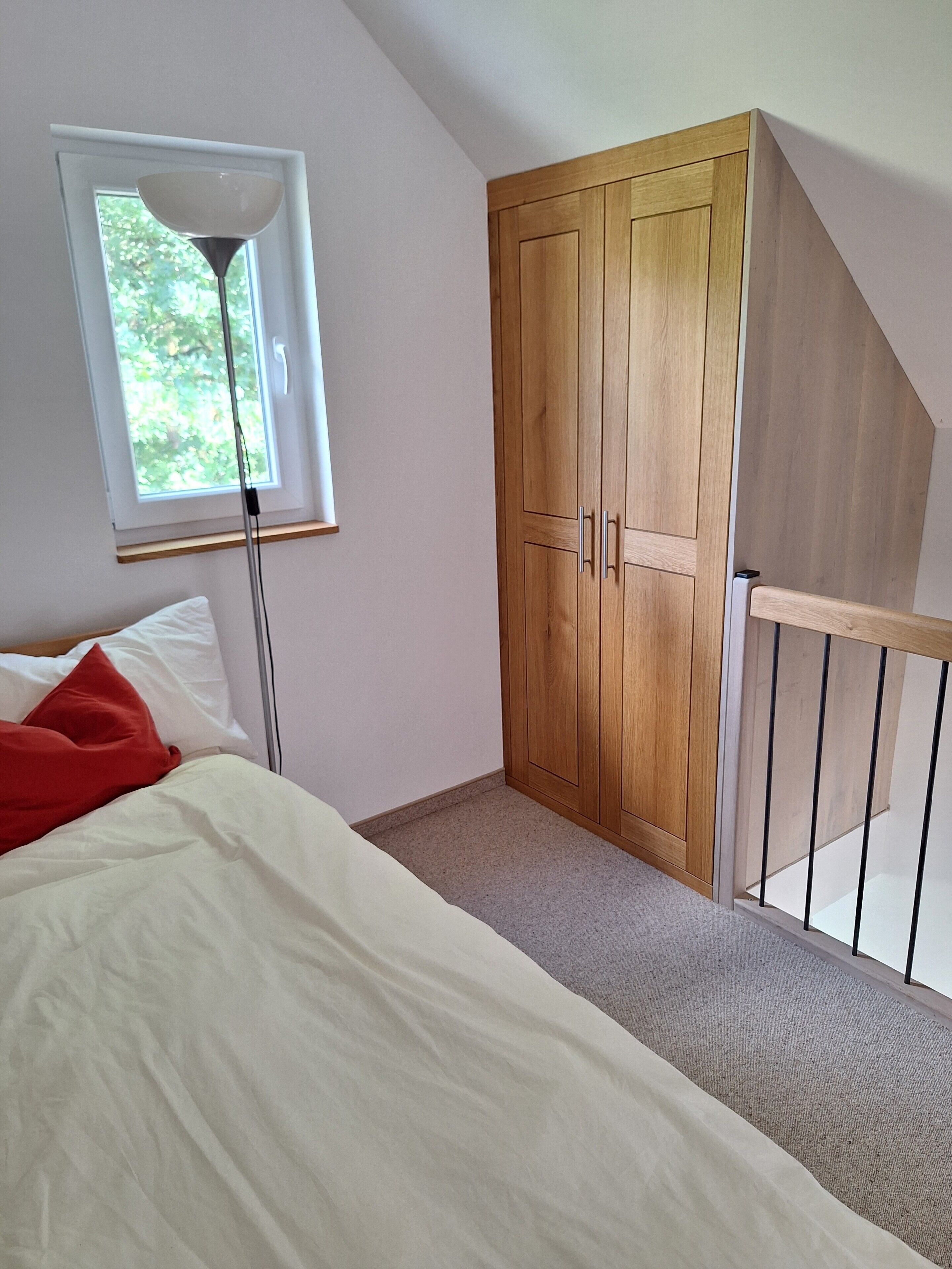 A bedroom with a bed, a window, a wooden wardrobe, and a staircase.
