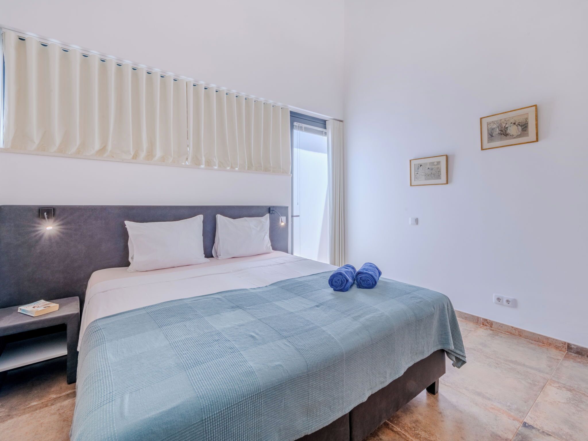 Een slaapkamer met een bed, een nachtkastje, een raam met gordijnen, en twee ingelijste foto's aan de muur.