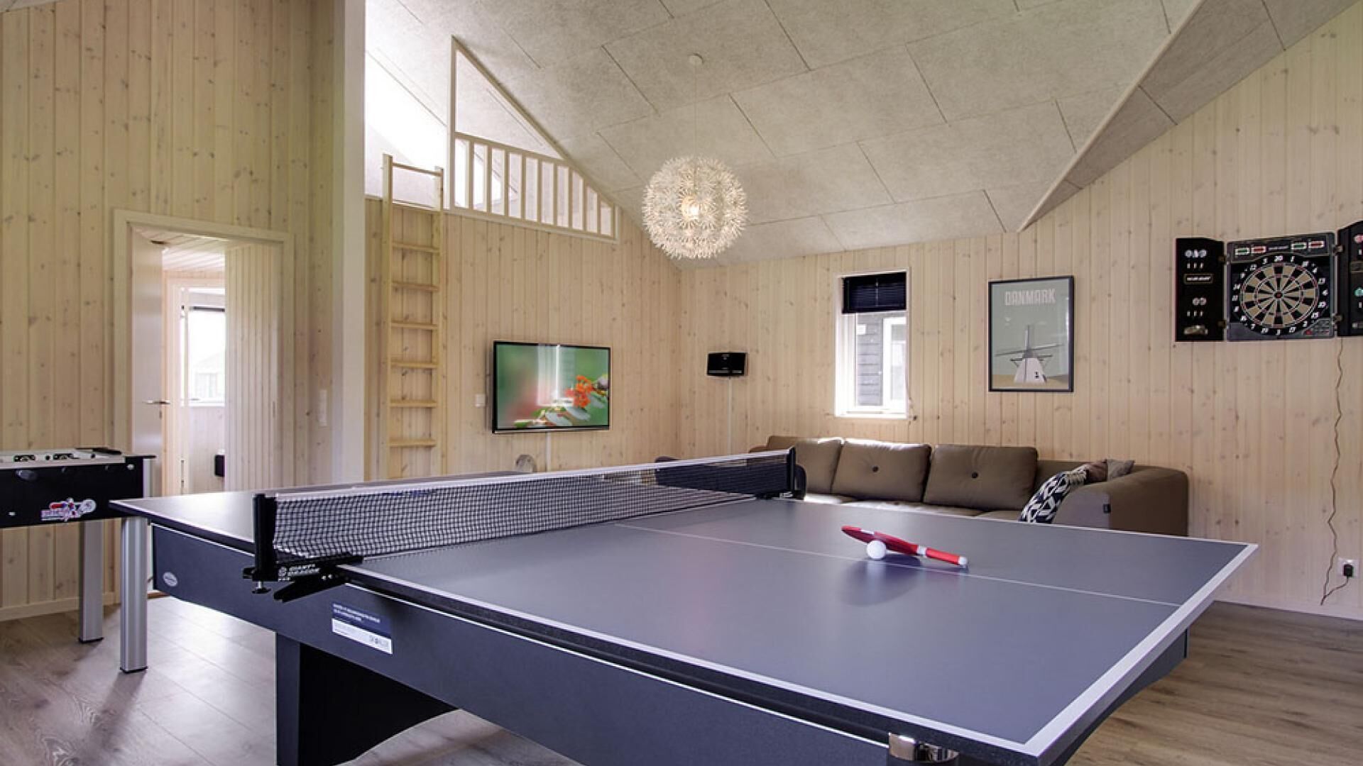 A ping pong table in a room with wooden walls, a loft bed, a TV, and dartboards.