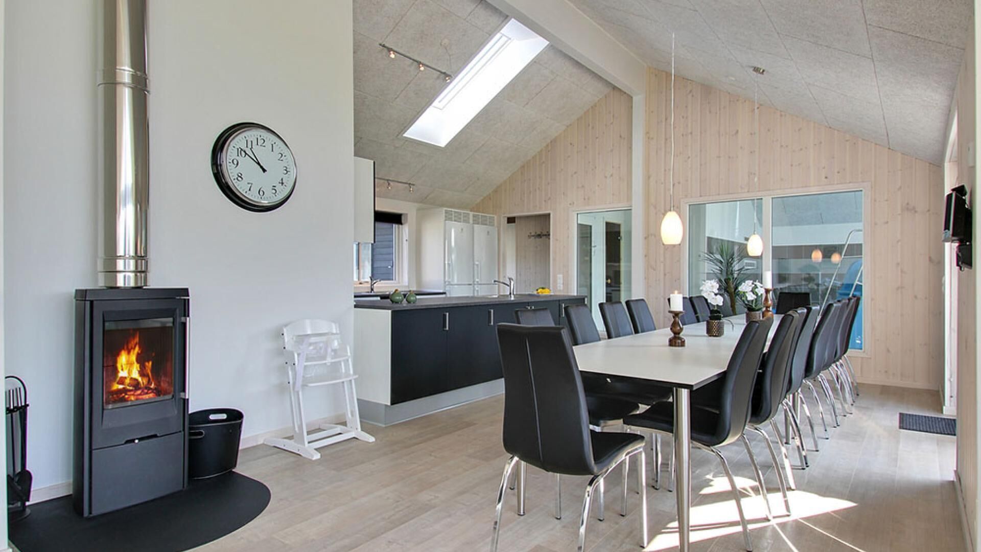 A modern kitchen with a wood-burning stove, a dining area with a long table and chairs, and a visible clock on the wall.