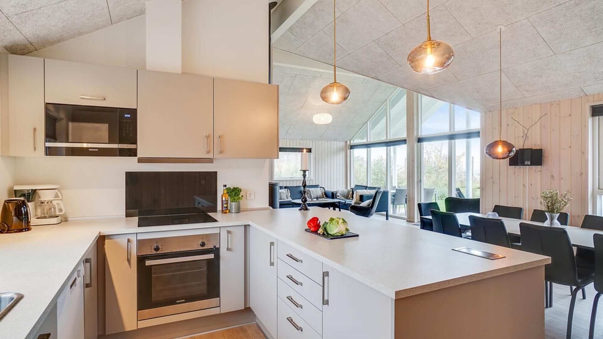 Eine moderne Küche mit weißen Schränken, einem eingebauten Ofen und einer großen Kochinsel mit Spüle. Es gibt einen Essbereich mit Tisch und Stühlen sowie ein großes Fenster für natürliches Licht.