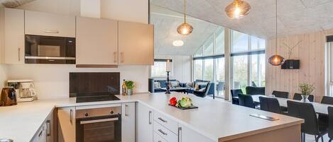 Eine moderne Küche mit weißen Schränken, einem eingebauten Ofen und einer großen Kochinsel mit Spüle. Es gibt einen Essbereich mit Tisch und Stühlen sowie ein großes Fenster für natürliches Licht.