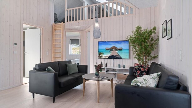 A modern living room with two black sofas, a coffee table, and a TV displaying a tropical scene. A wooden ladder and a balcony are visible.
