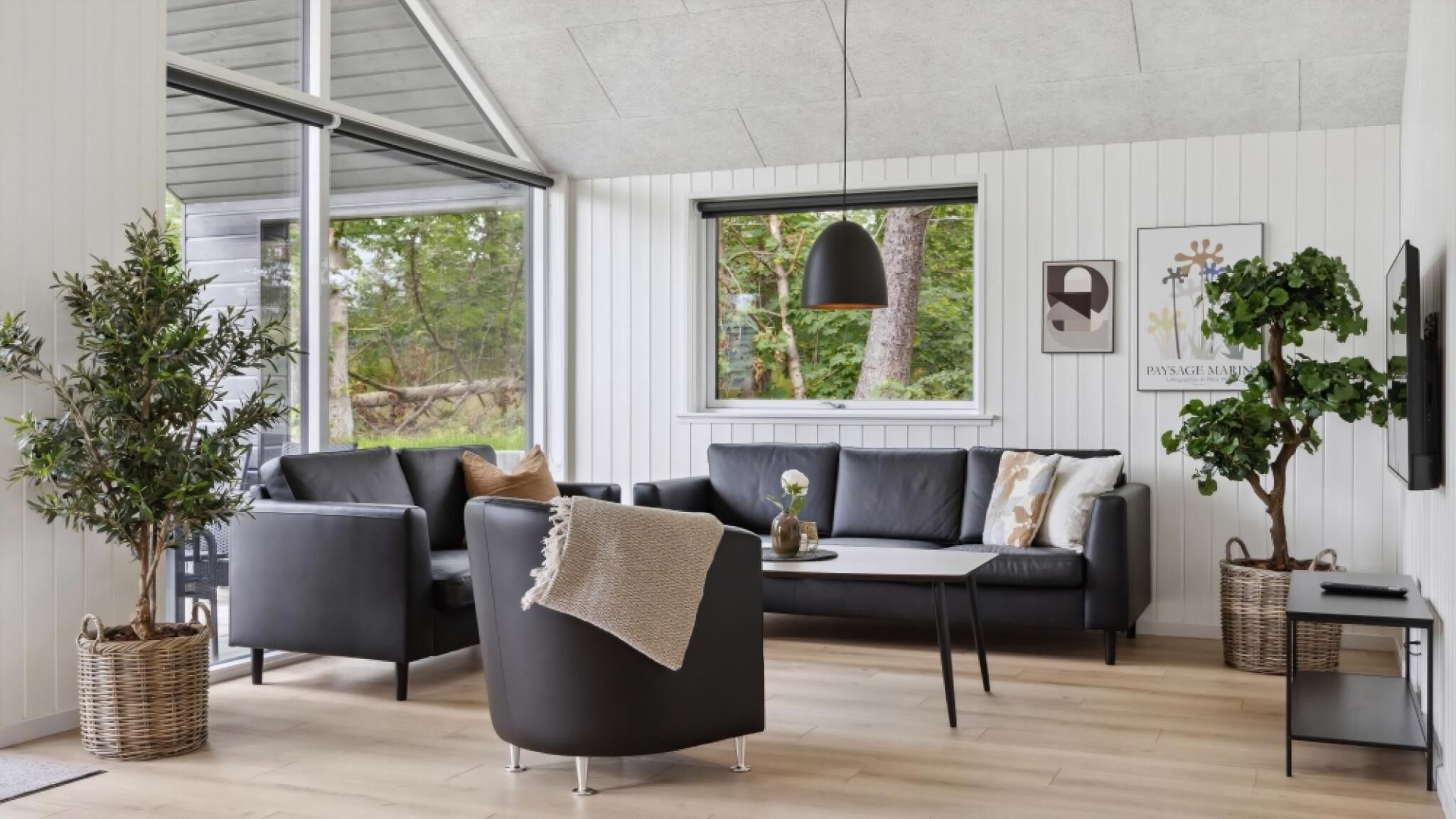 Een moderne woonkamer met een zwarte bank, twee fauteuils, een salontafel en een groot raam met uitzicht op bomen. Er staan twee potplanten en er hangt een lamp.
