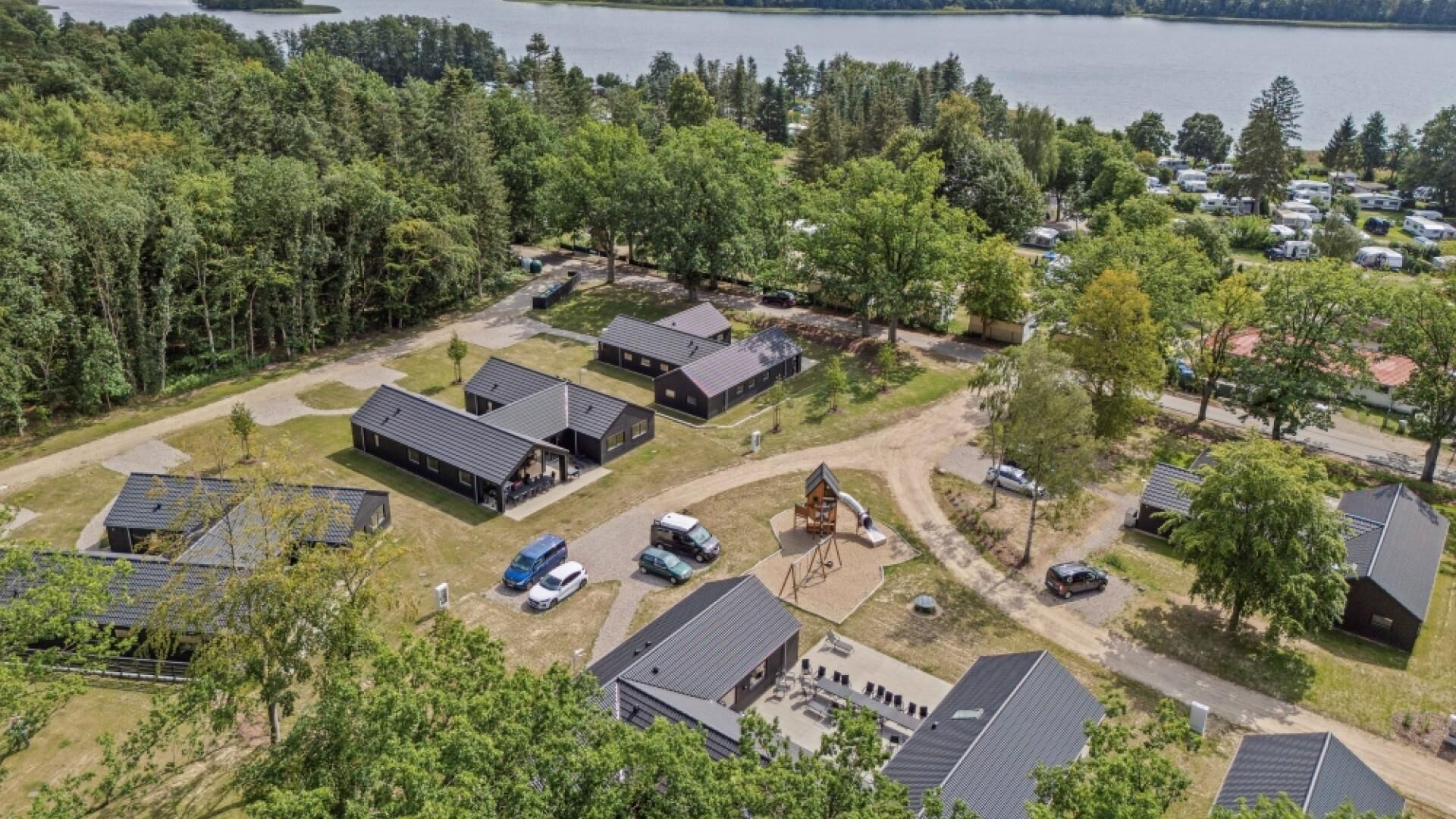 Vista aérea de un camping con varias cabañas, un parque infantil y un lago al fondo.