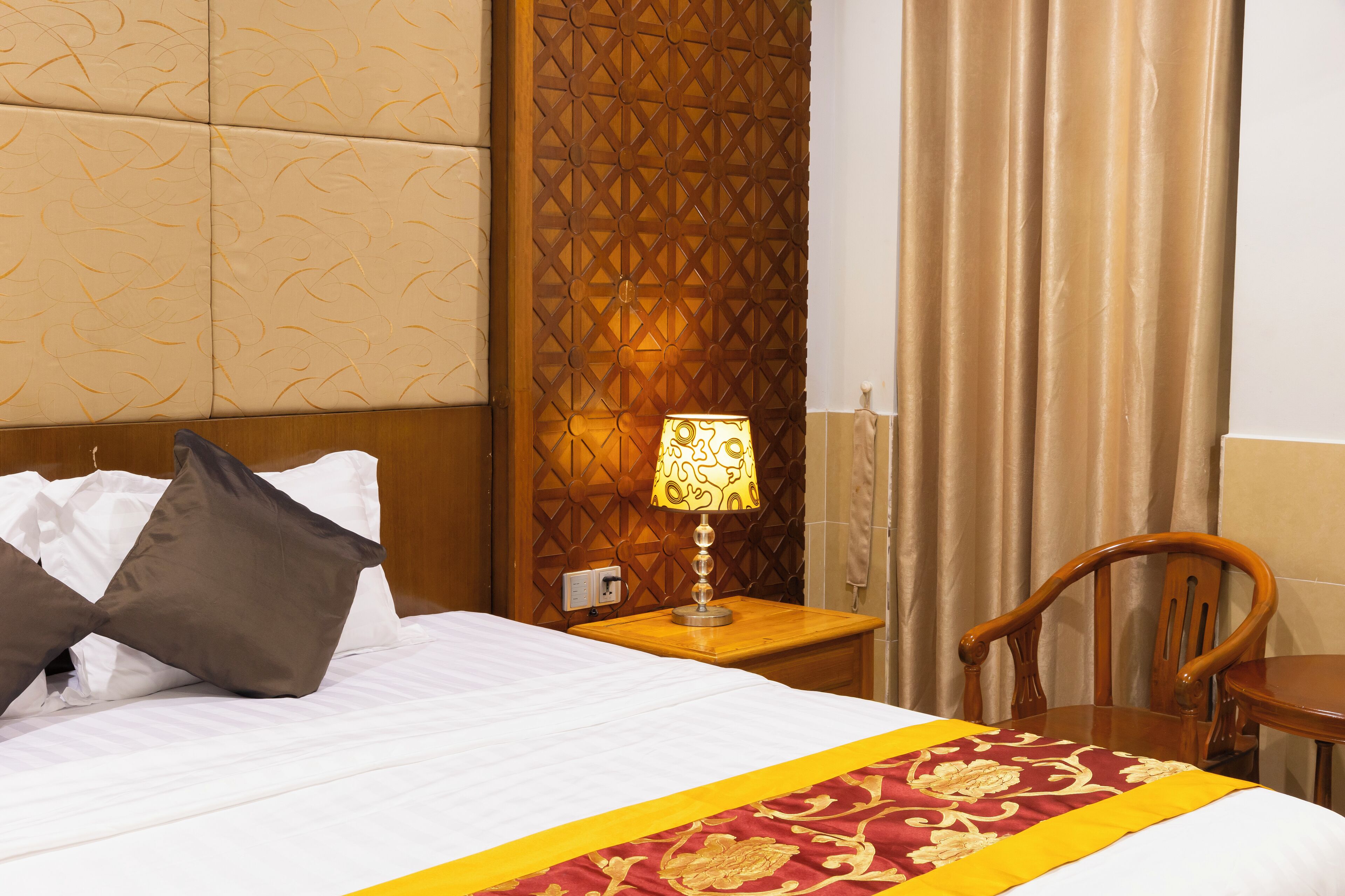 A hotel room with a bed, pillows, a nightstand with a lamp, a wooden chair, and a curtain.