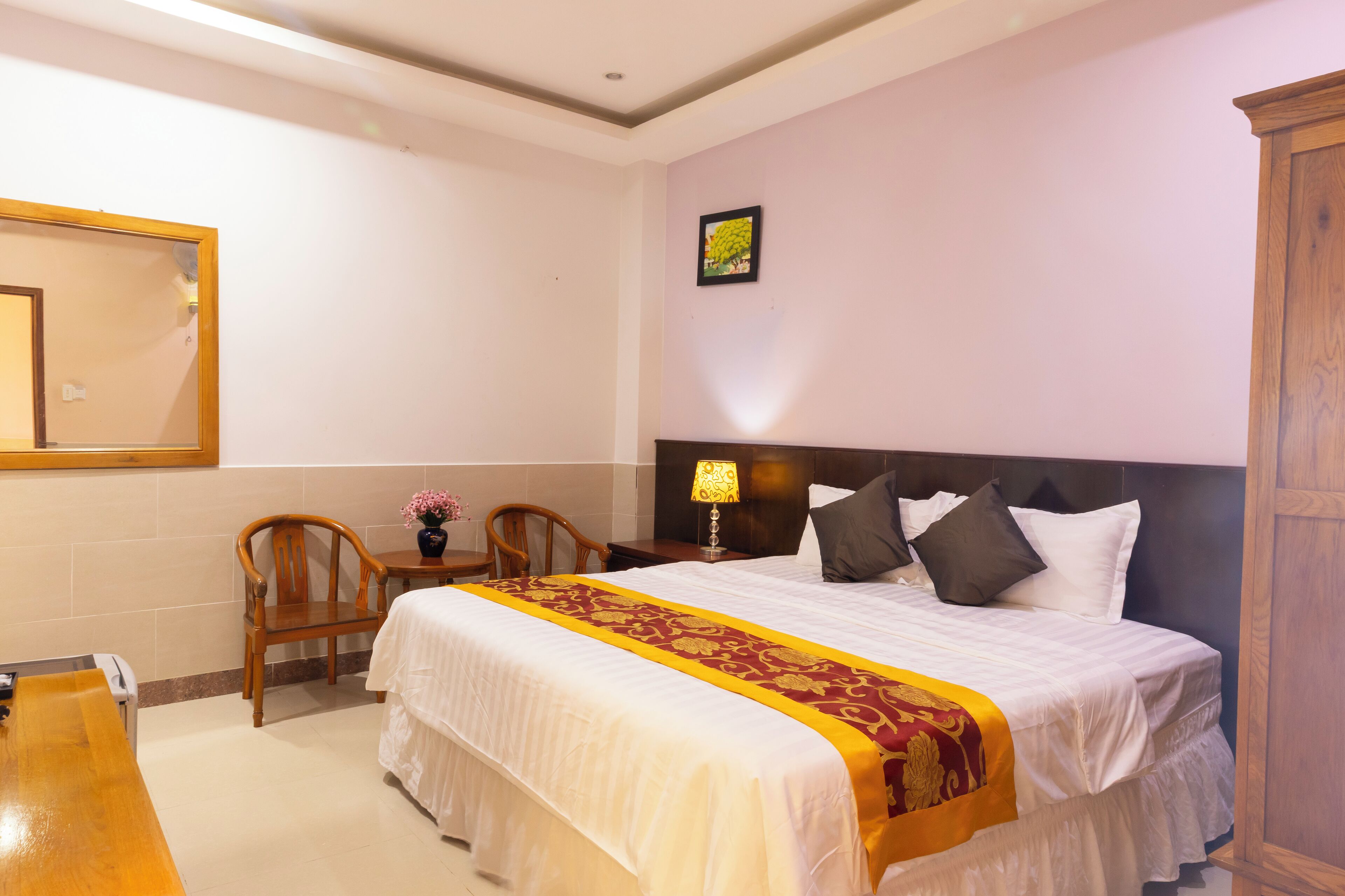 A hotel room with a large bed, a wooden nightstand, a chair, a desk, and a mirror on the wall.