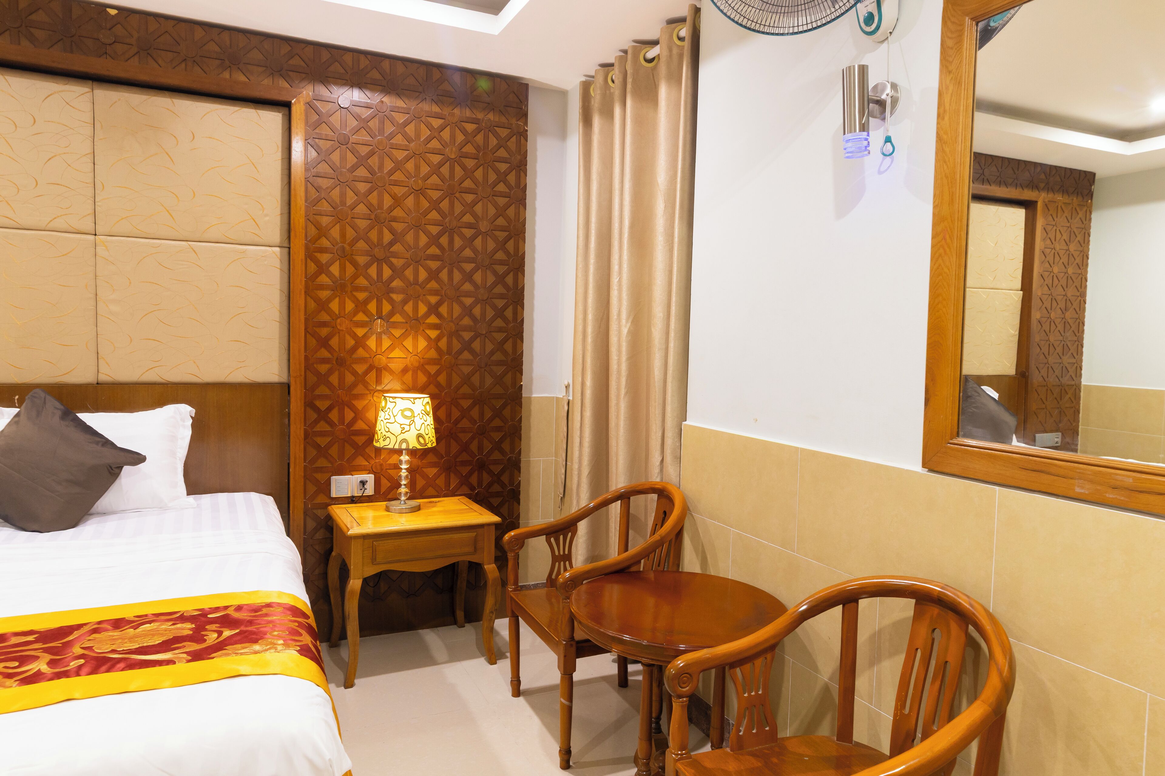A hotel room with a bed, a desk with a lamp, two wooden chairs, and a mirror on the wall.