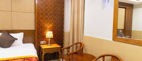 A hotel room with a bed, a desk with a lamp, two wooden chairs, and a mirror on the wall.