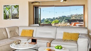 Una sala de estar moderna con techo de madera, un ventanal con vista a la ciudad, un sofá color beige con cojines amarillos y dos mesas de centro blancas.