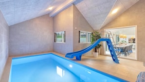 Una piscina interior con un tobogán azul, un comedor con mesa y sillas, y un gran ventanal con cortinas.