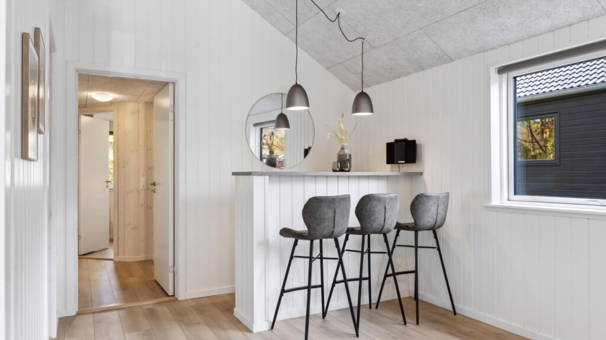 Una cocina moderna con encimera blanca, tres taburetes de bar, luces colgantes y un amplio ventanal con vistas.