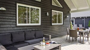 Une maison en bois aux couleurs sombres, avec un canapé noir et une table dressée, sur laquelle on peut voir une bouteille, des verres et une assiette de nourriture.