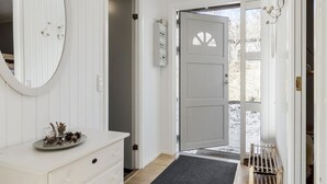 Un intérieur blanc avec un grand miroir, une commode et une porte donnant sur l’extérieur.
