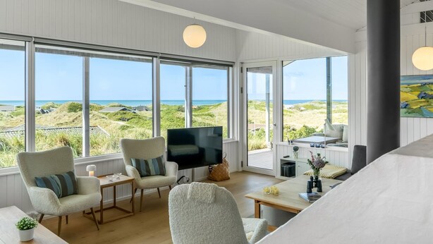 Ein großzügiges Wohnzimmer mit großen Fenstern und Blick auf Strand und Dünen.