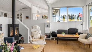 Ein gemütliches Wohnzimmer mit Kaminofen, einer bequemen Couch, einem Holztisch und Blick ins Freie.