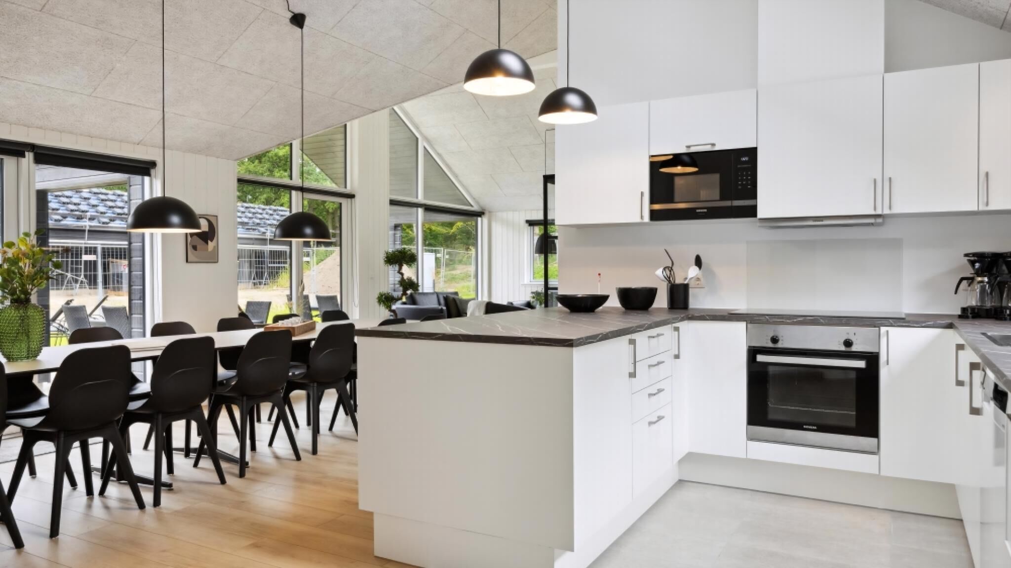 Una cocina moderna con armarios blancos, una isla central, lámparas colgantes negras y vistas al exterior a través de ventanas amplias.