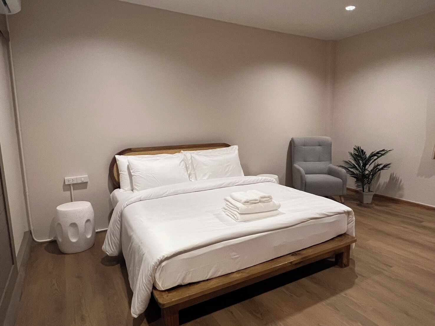 A modern bedroom with a wooden bed frame, white bedding, a grey armchair, and a potted plant.