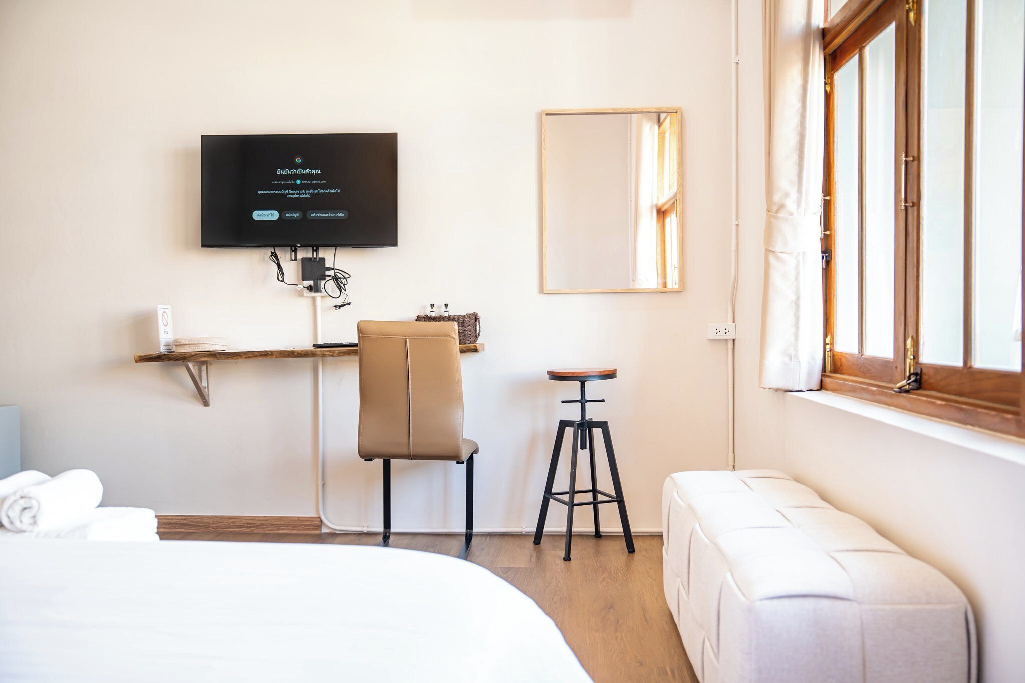 Una habitación de hotel con cama, escritorio y silla, televisor montado en la pared, ventana con cortinas y mesita de noche con toallas.
