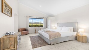 Une chambre à coucher avec un grand lit, deux tables de chevet, une chaise, une armoire en bois et une fenêtre donnant sur un paysage verdoyant.